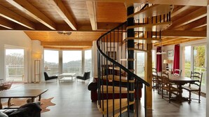 Amplia sala de estar con escalera de madera, ventanas grandes y zona de comedor.