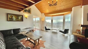 Un salón amplio con techo alto, vigas de madera, un ventanal con vistas al exterior, un sofá de cuero negro, una mesa de centro y una araña.