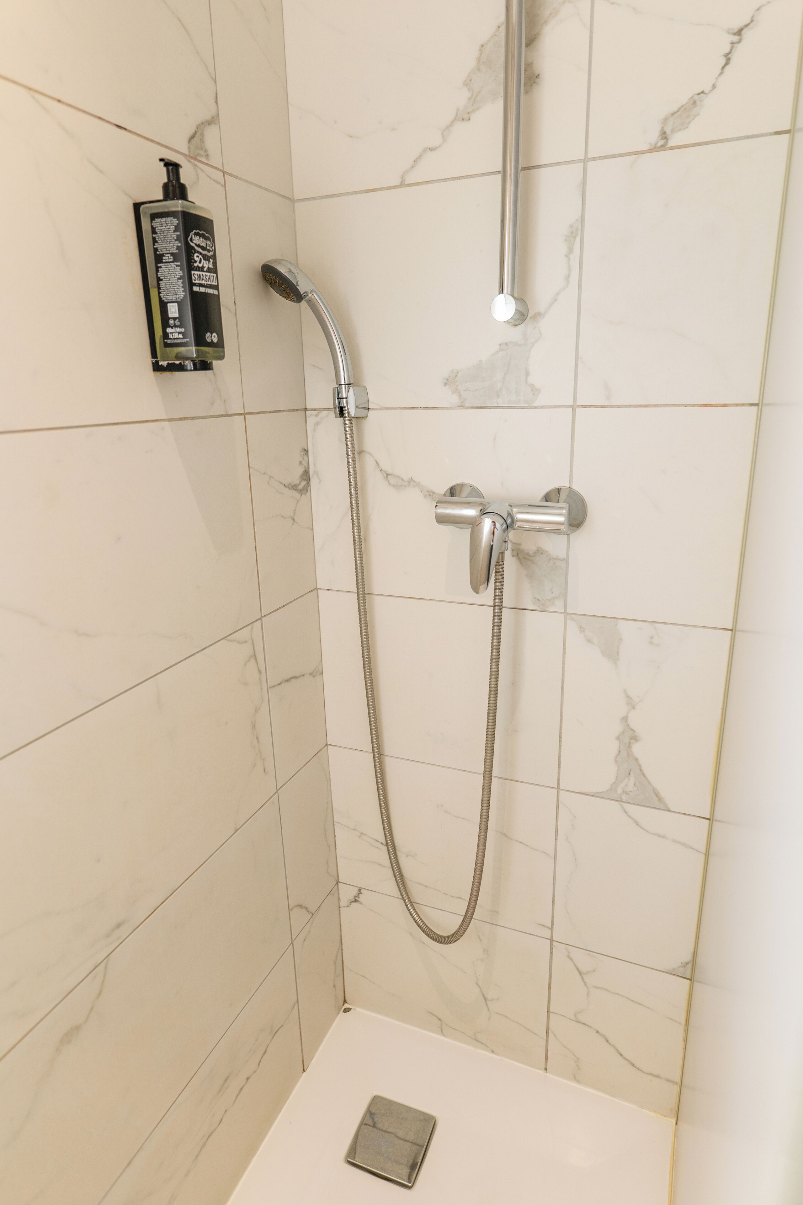 Doccia con soffione a mano e rubinetto montato a parete, erogatore di sapone e piccolo vassoio metallico alla base.