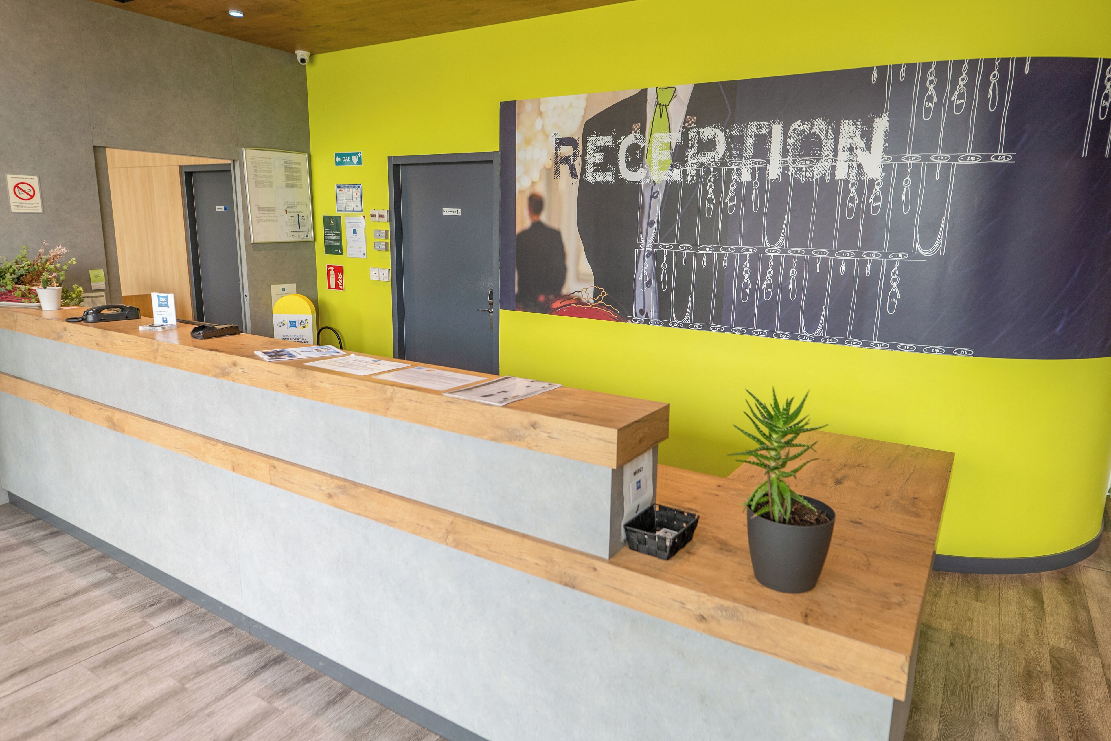 Un mostrador de recepción con encimera de madera y base gris, una planta en maceta y un cartel grande que dice 'RECEPTION'.