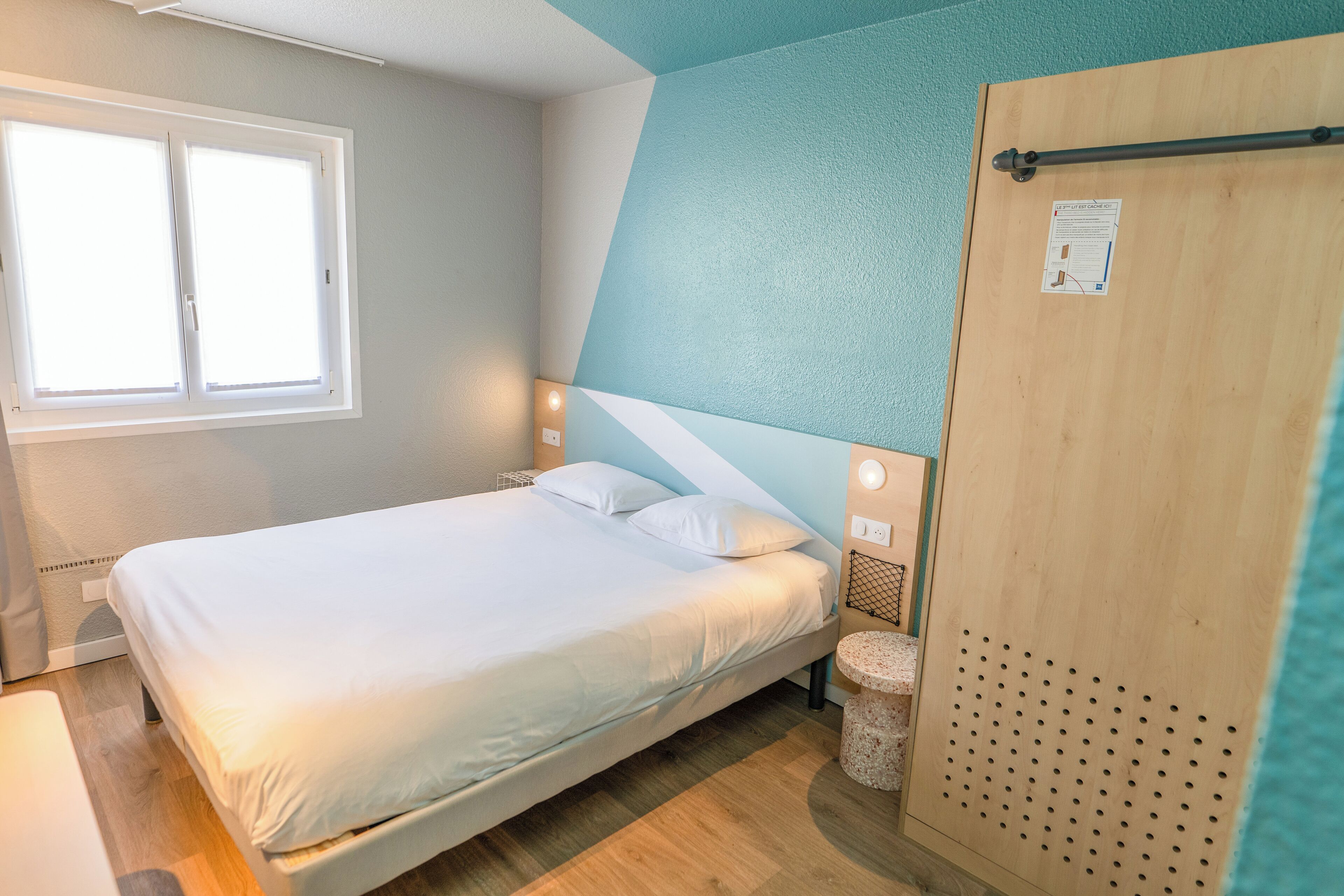 Una cama bien hecha en una habitación con ventana, una puerta de madera y una pequeña mesa redonda.