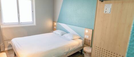 A neatly made bed in a room with a window, a wooden door, and a small round table.
