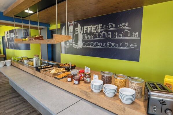 A buffet counter with various food items and condiments, a chalkboard with food illustrations, and a toaster. - Ibis Budget Brest Sud Plougastel - Hôtel Rénové (Plougastel-Daoulas)