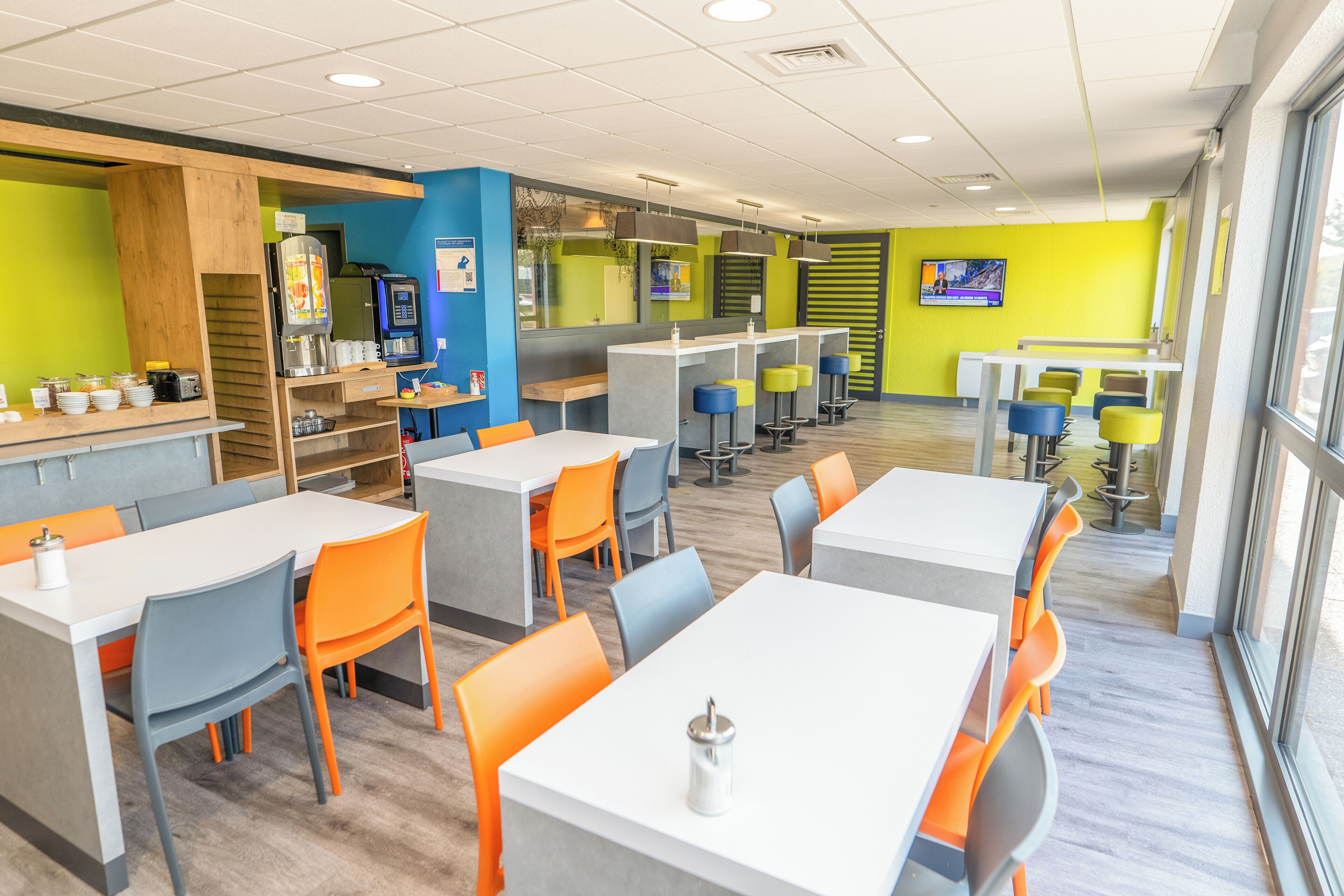 Een moderne cafetaria met meerdere tafels en stoelen, een koffieapparaat en een televisiescherm.