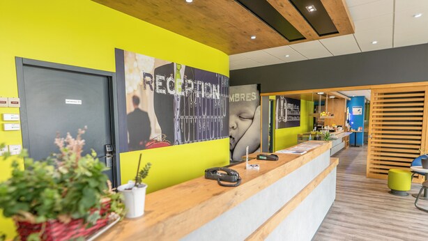 A reception area with a wooden reception desk, a large reception sign, and a potted plant. - Ibis Budget Brest Sud Plougastel - Hôtel Rénové (Plougastel-Daoulas)