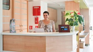 Een receptiebalie met een vrouw erachter, een waterdispenser, een brandblusser en een kamerplant.