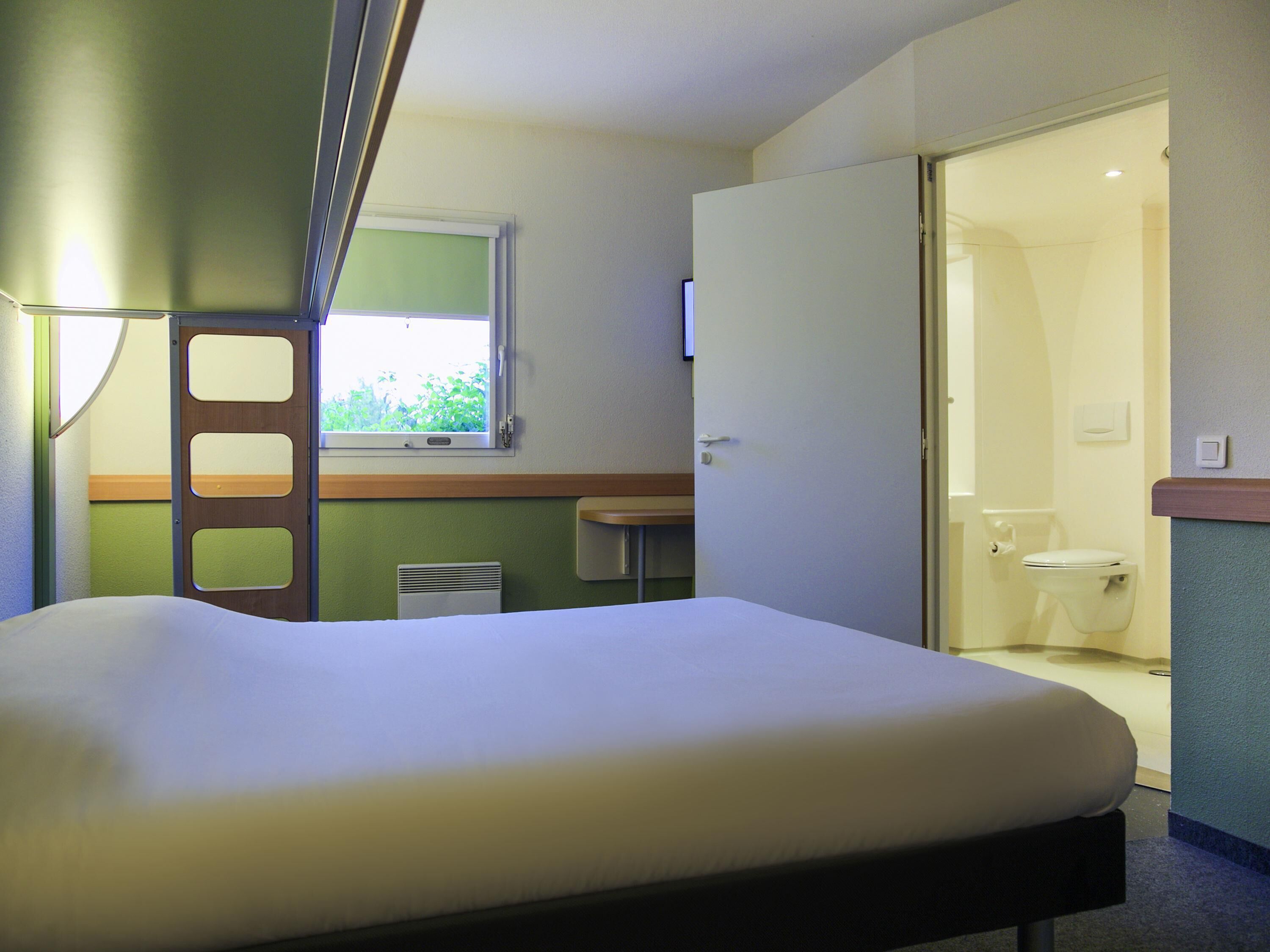 Una cama individual en una habitación con ventana, una puerta que da a un baño y una mesita.
