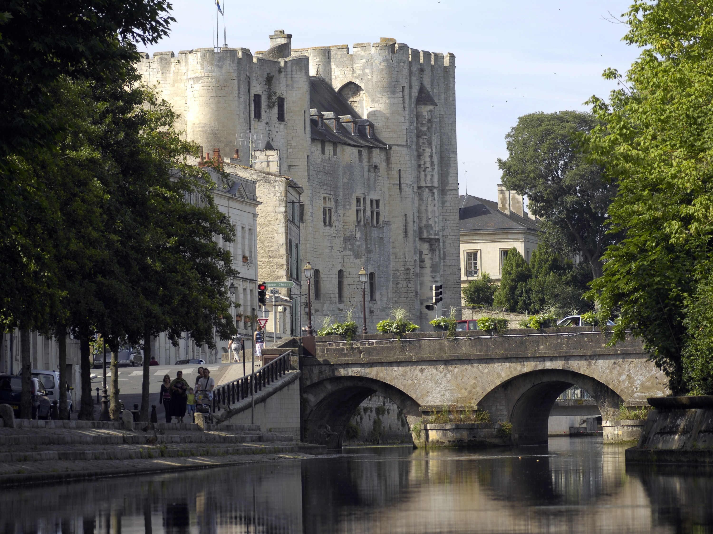 Un pont de pierre enjambant une rivière paisible, un château historique en arrière-plan et des piétons sur le pont.
