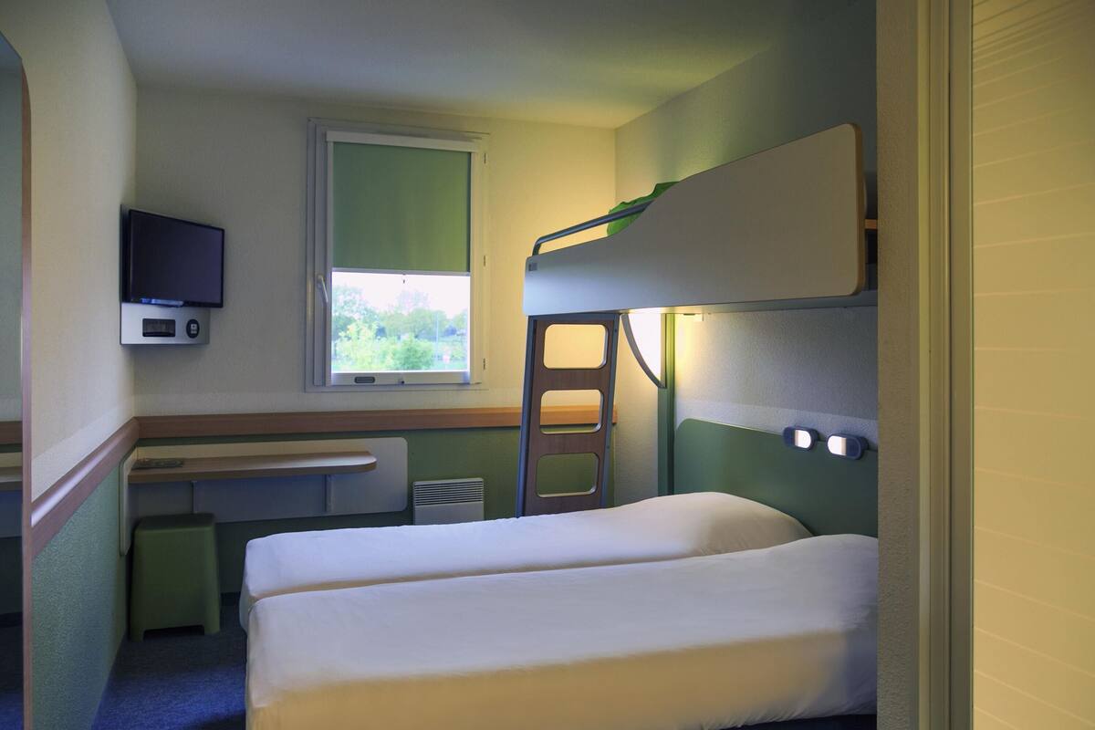 Een kleine, compacte kamer met stapelbedden, een bureau, een televisie en een raam met uitzicht op groen.