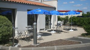 Een terras met blauwe parasols, tafels en stoelen voor een gebouw met een betegeld dak.