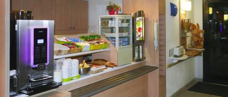 Zona de desayuno autoservicio con máquina de café, vitrina refrigerada y diversas opciones de alimentos.