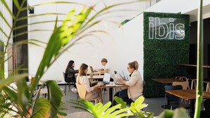 A modern indoor space with a green wall and the word "ibis" on it. There are people sitting at tables with laptops, suggesting a co-working area. - ibis Puerto General San Martin (Puerto General San Martín)