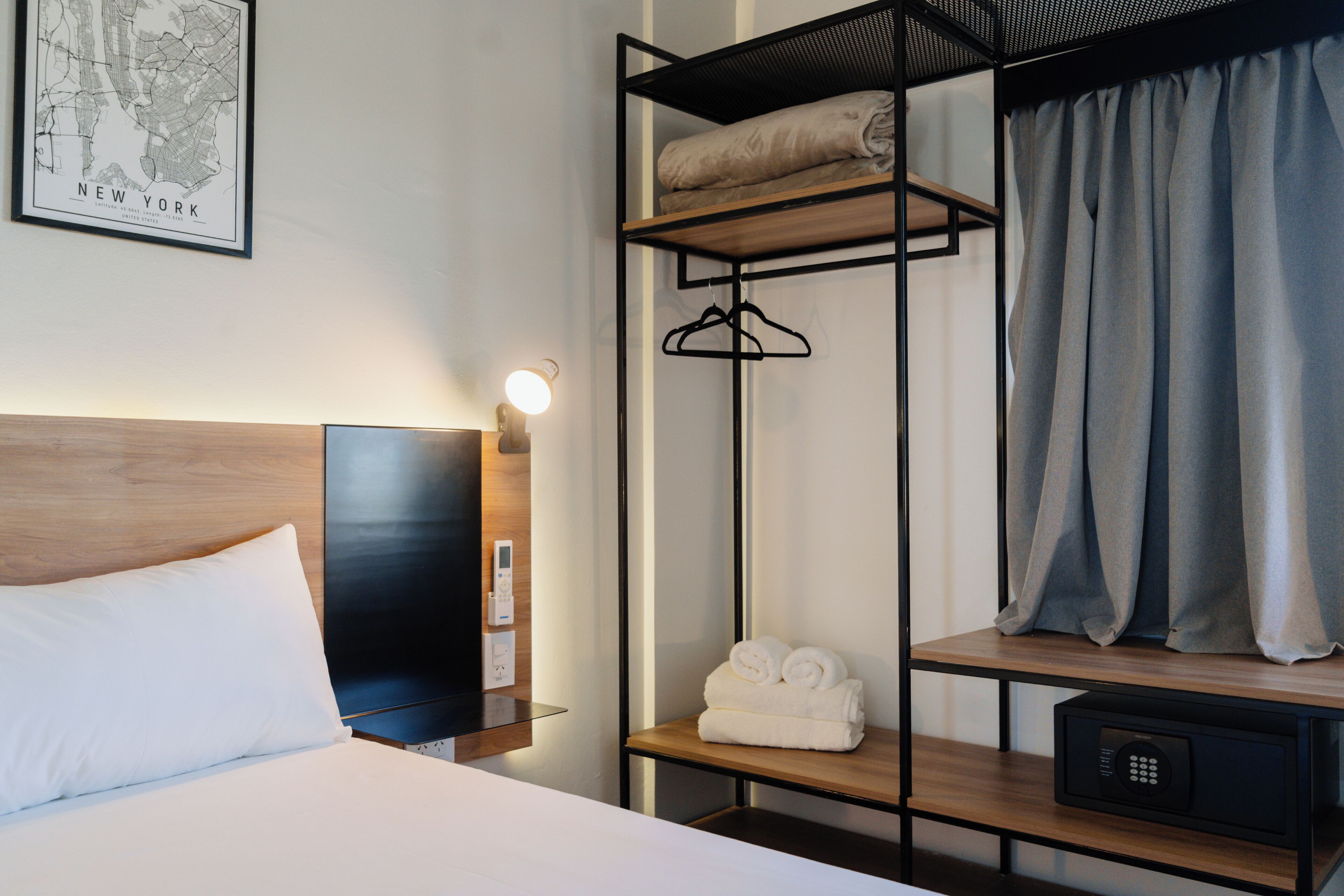 Habitación de hotel con cama, cabecera de madera, televisor montado en la pared, mapa de Nueva York, estante con toallas, perchero y cortina.