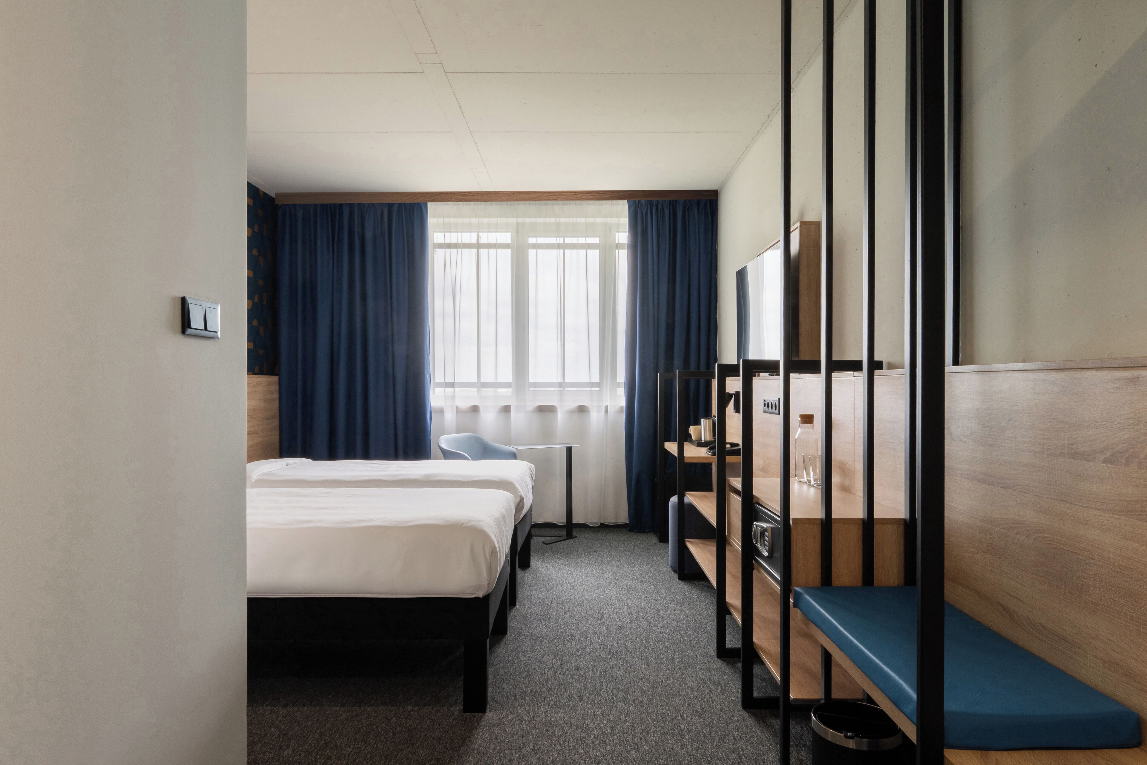 A hotel room with two beds, a desk, a chair, a small table, a mirror, and a window with curtains.