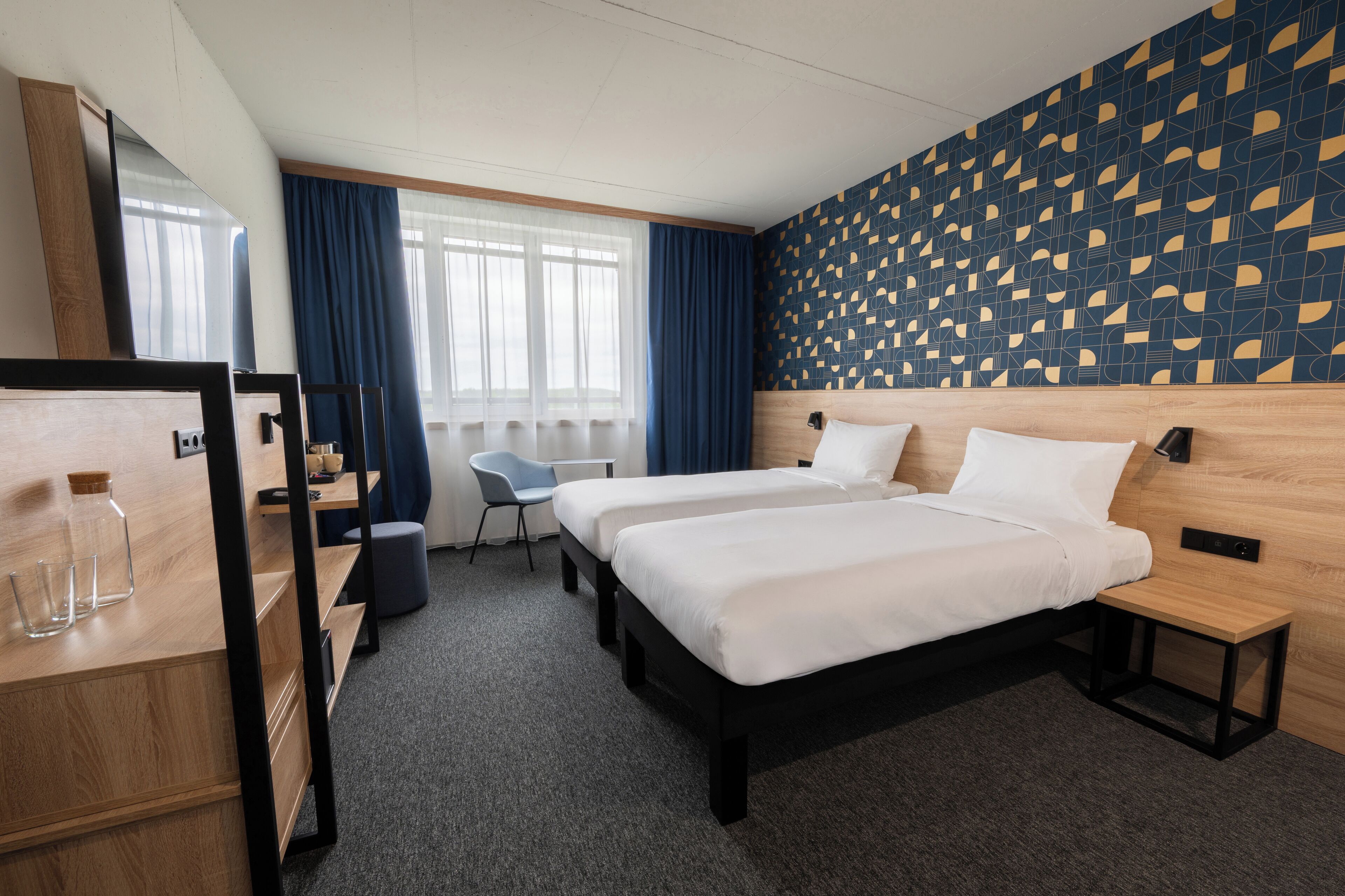 A hotel room with two beds, a desk, a chair, and a large window with curtains.