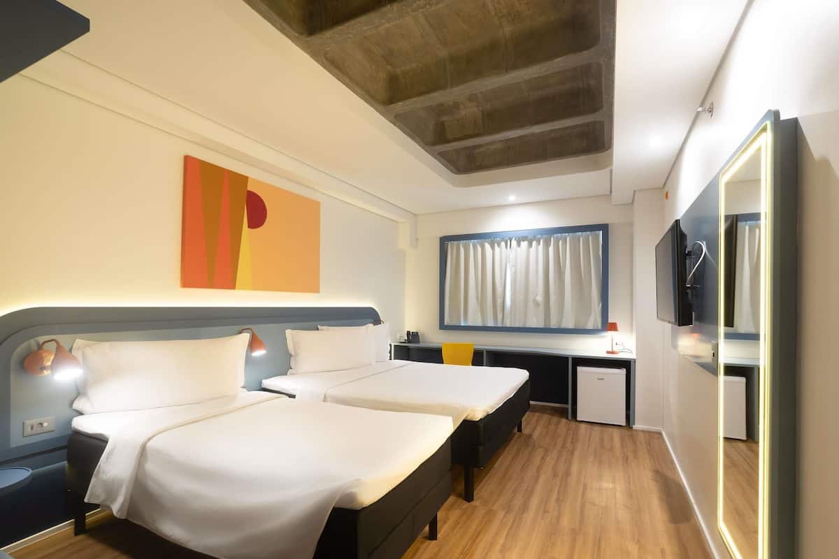 A hotel room with two beds, a desk, a television, and a large window with curtains.