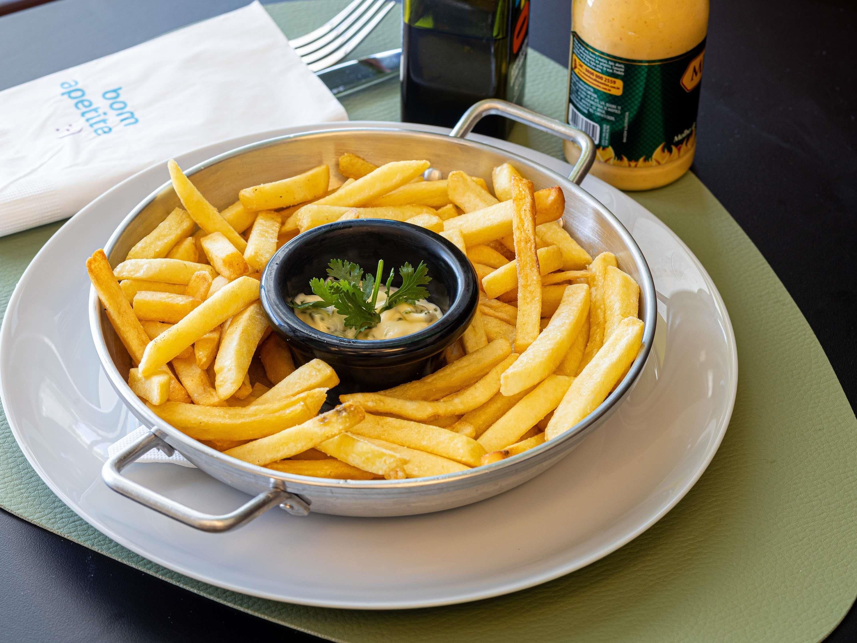 Um prato de batatas fritas com molho para acompanhar e uma pequena tigela de molho, colocado sobre uma mesa com um guardanapo e uma garrafa ao fundo.