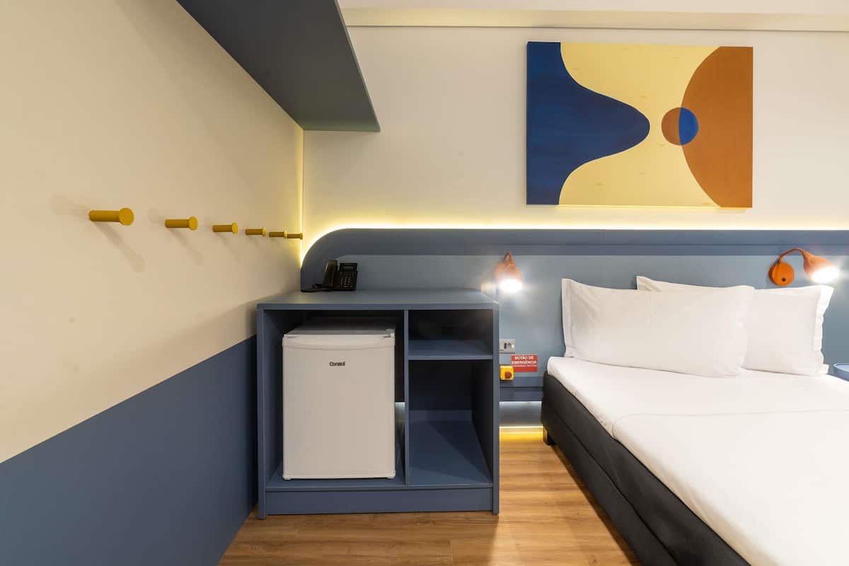 A hotel room with a bed, a small fridge, a bedside table, a wall-mounted phone, and a painting.