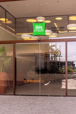 La entrada a ibis Styles con puerta de cristal, un letrero verde y lámparas colgantes.