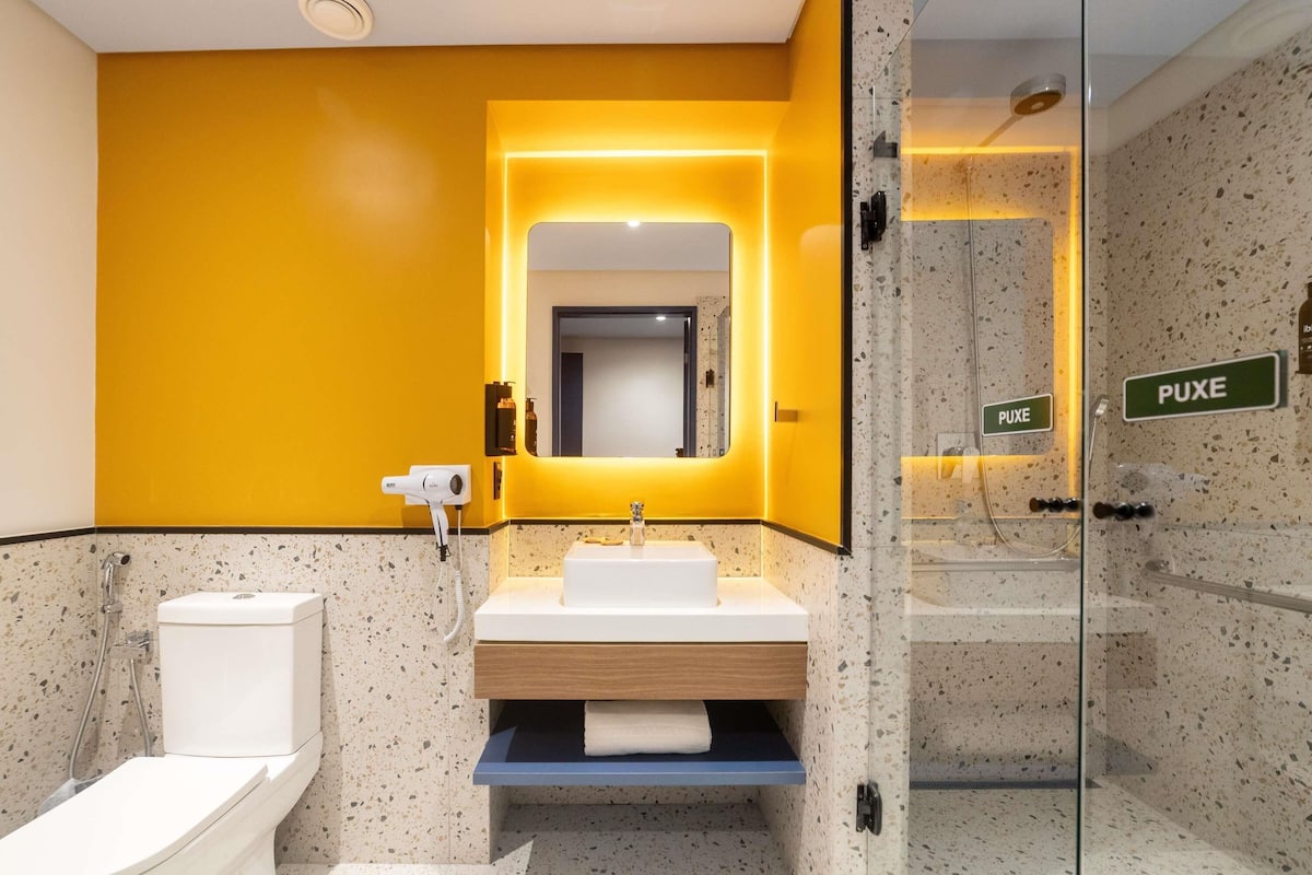 Un baño moderno con una pared de acento amarilla, cerramiento de ducha de vidrio y un letrero visible que dice "PUXE".