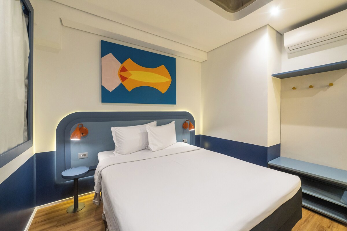 A hotel room with a bed, a small table, a wall-mounted lamp, and a colorful abstract painting.