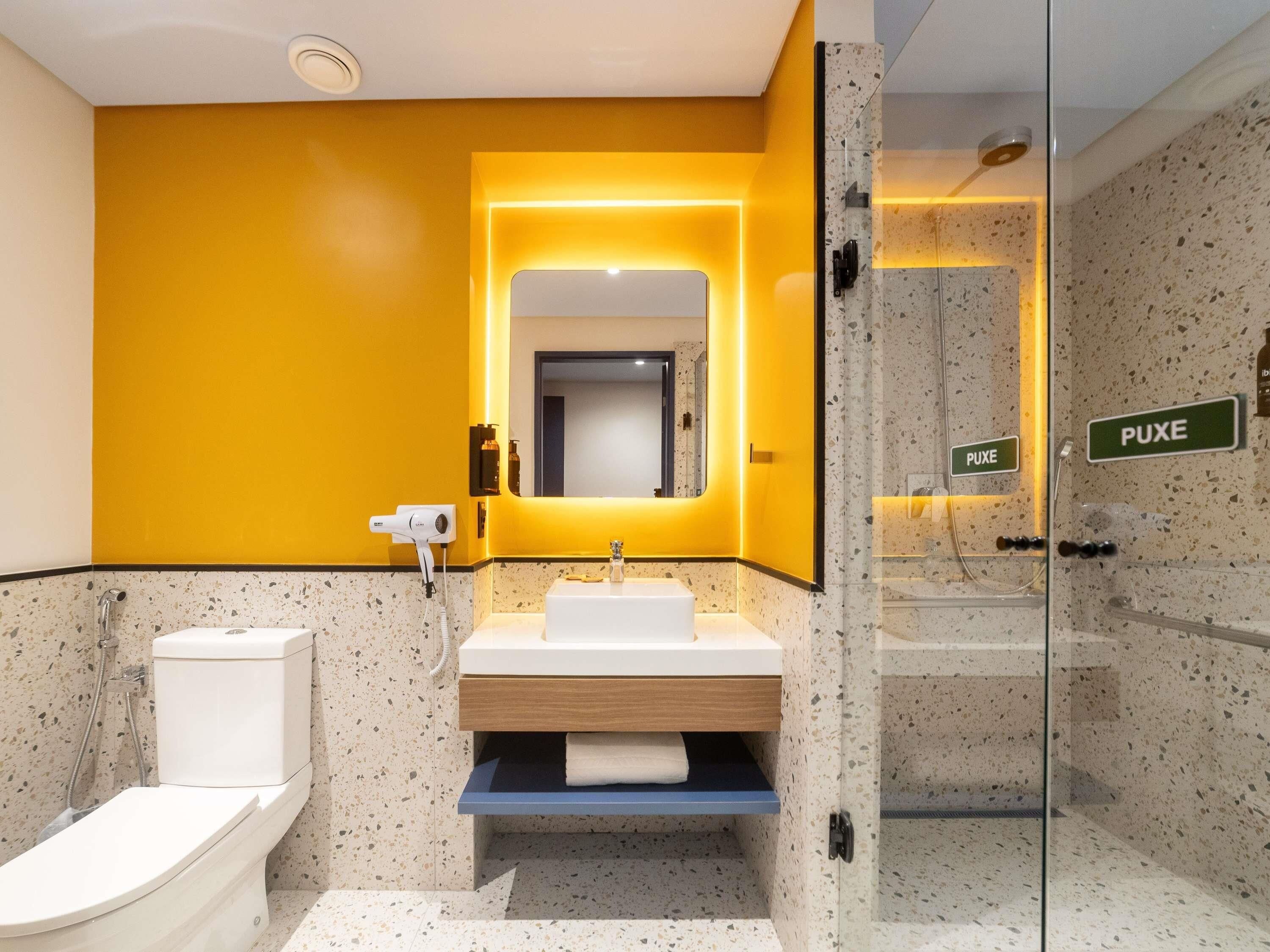 Un baño moderno con una pared de acento amarilla, cerramiento de ducha de vidrio y un letrero visible que dice "PUXE".