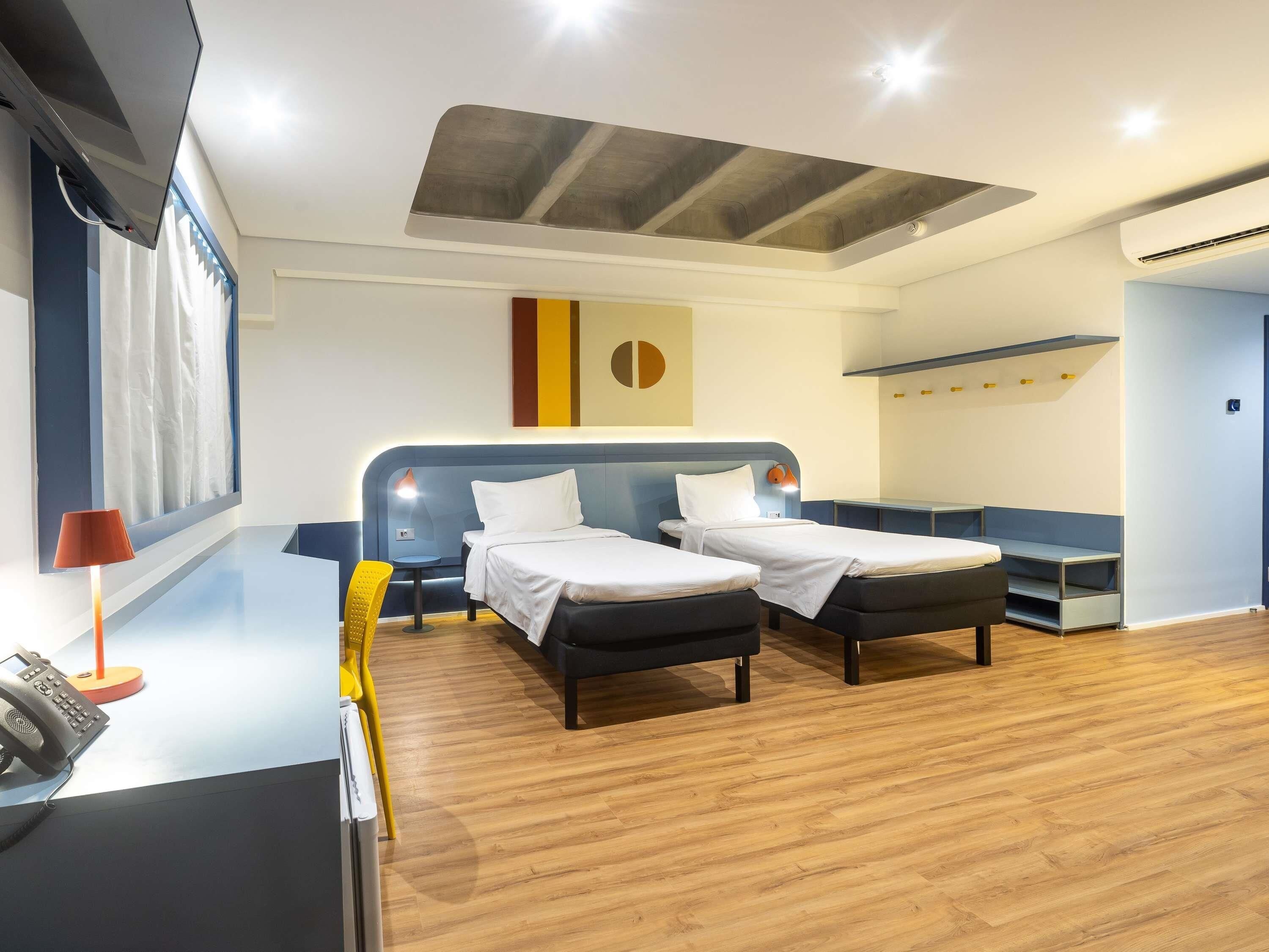 Una habitación de hotel moderna con dos camas individuales, un escritorio con teléfono y un ventanal con cortinas.