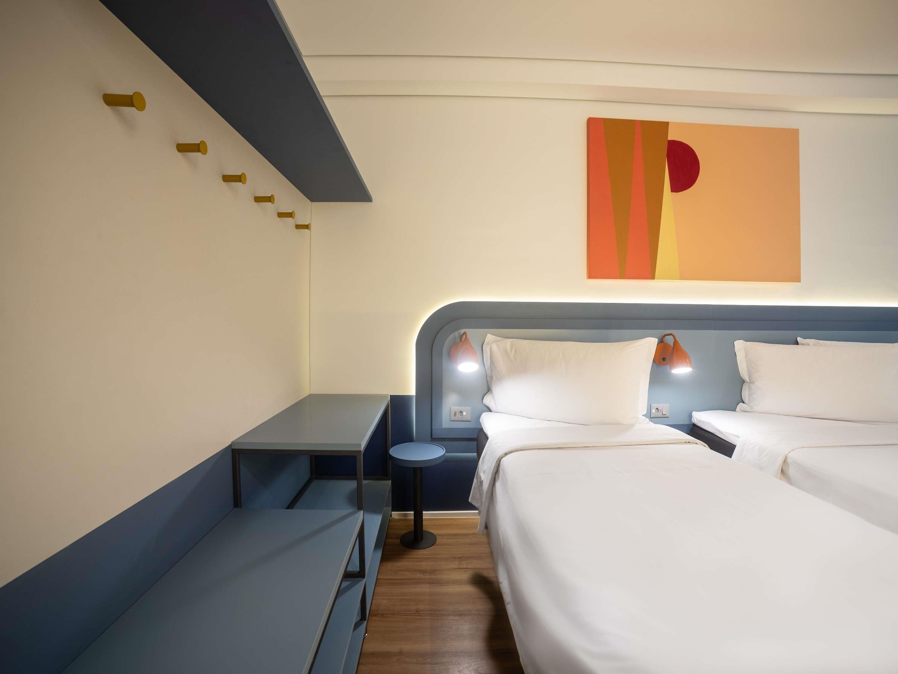 Quarto de hotel com cama, mesas de cabeceira, escrivaninha e uma obra de arte na parede.
