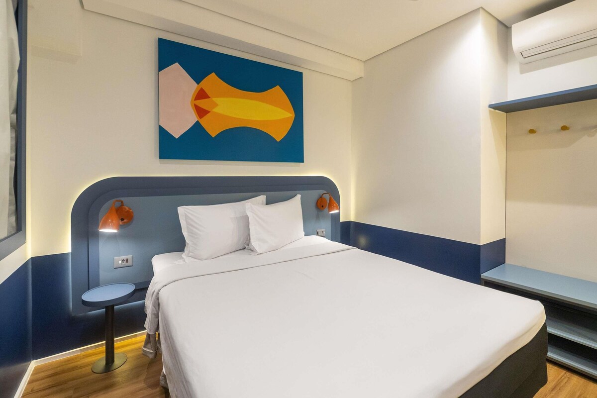 A hotel room with a bed, a bedside table, an air conditioning unit, and a wall-mounted artwork.