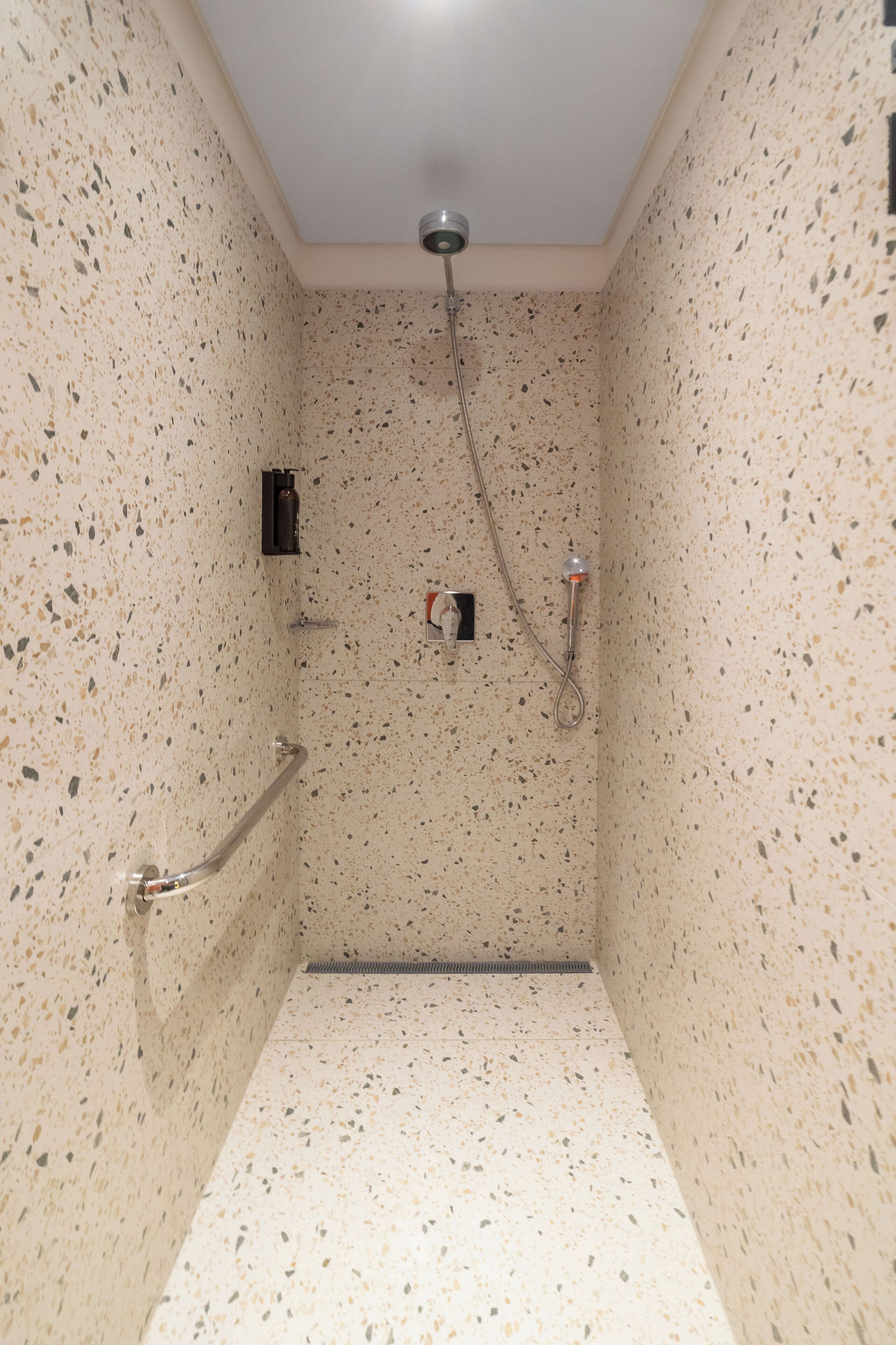A small, enclosed shower area with a handheld showerhead, a handheld faucet, and a wall-mounted soap dispenser.