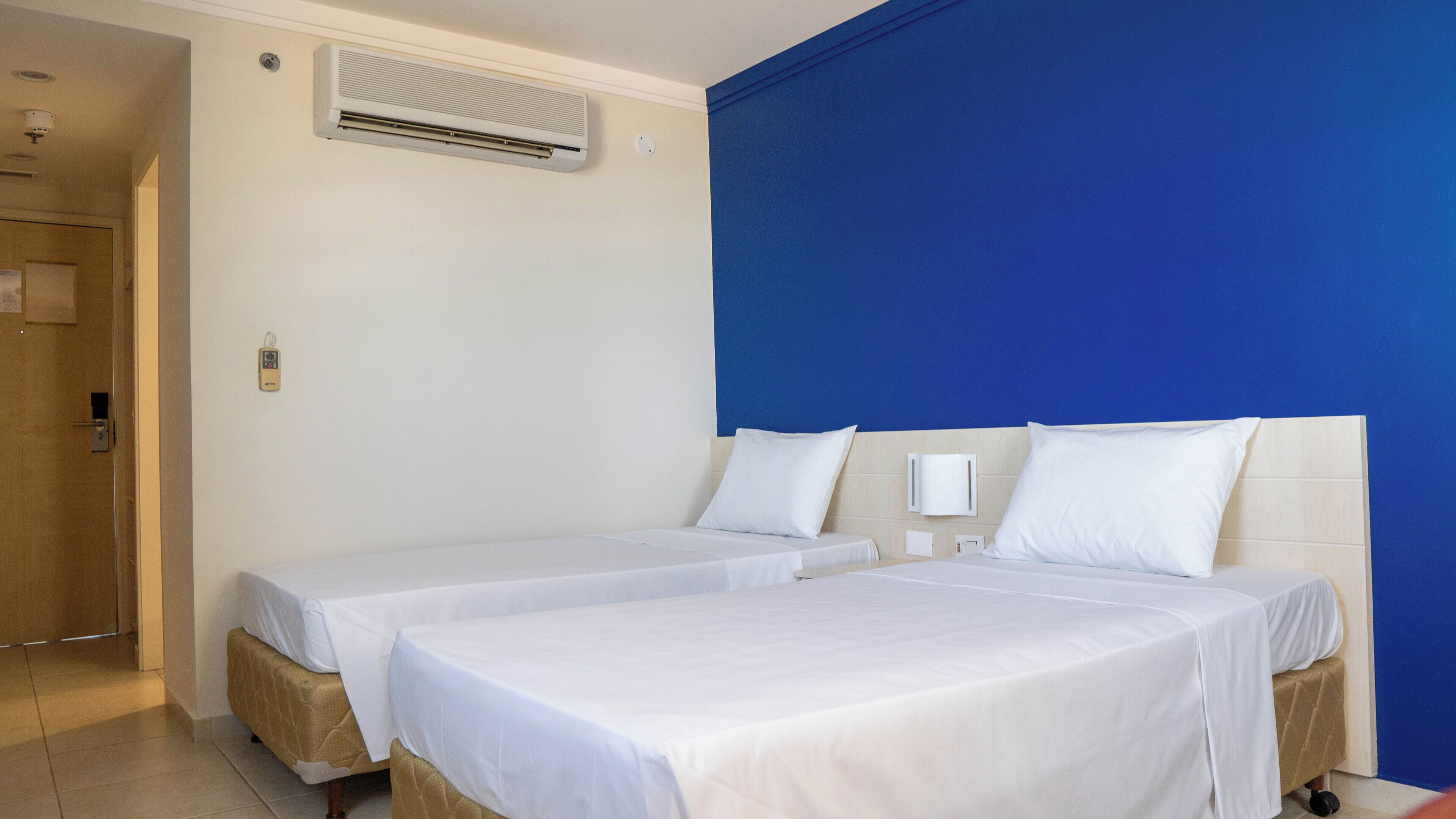 A hotel room with two beds, a blue accent wall, an air conditioning unit, and a wooden door.