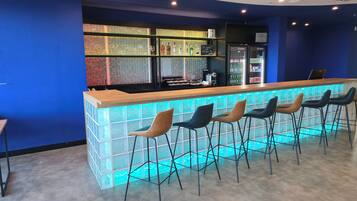 A bar area with a wooden counter and bar stools. There are shelves with bottles and glasses behind the counter.