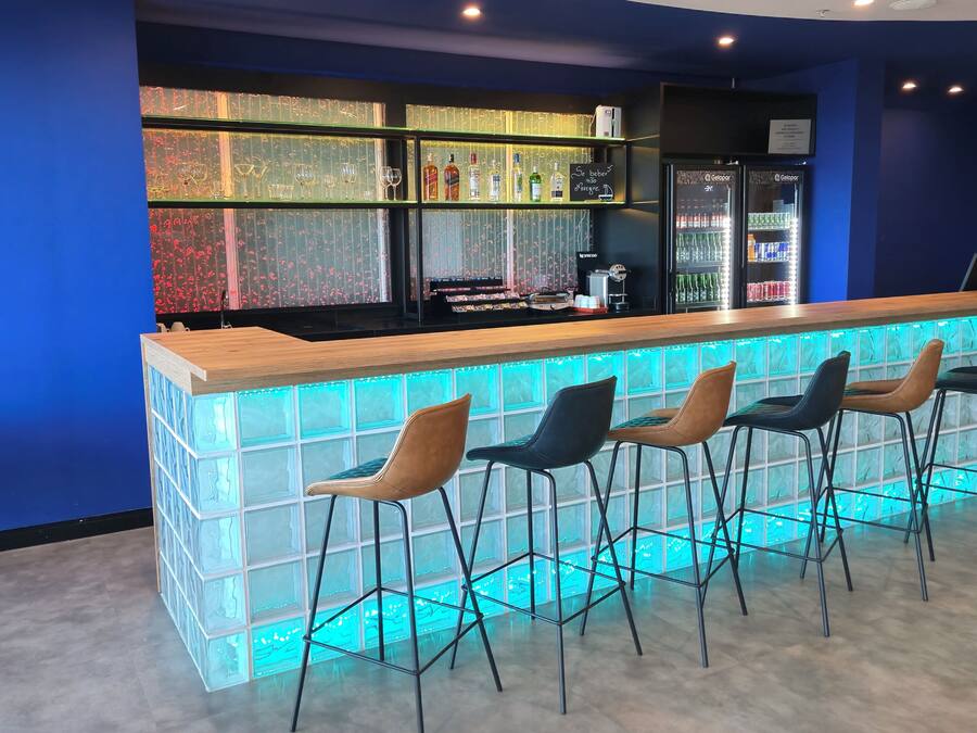 A bar area with a wooden counter and bar stools. There are shelves with bottles and glasses behind the counter.