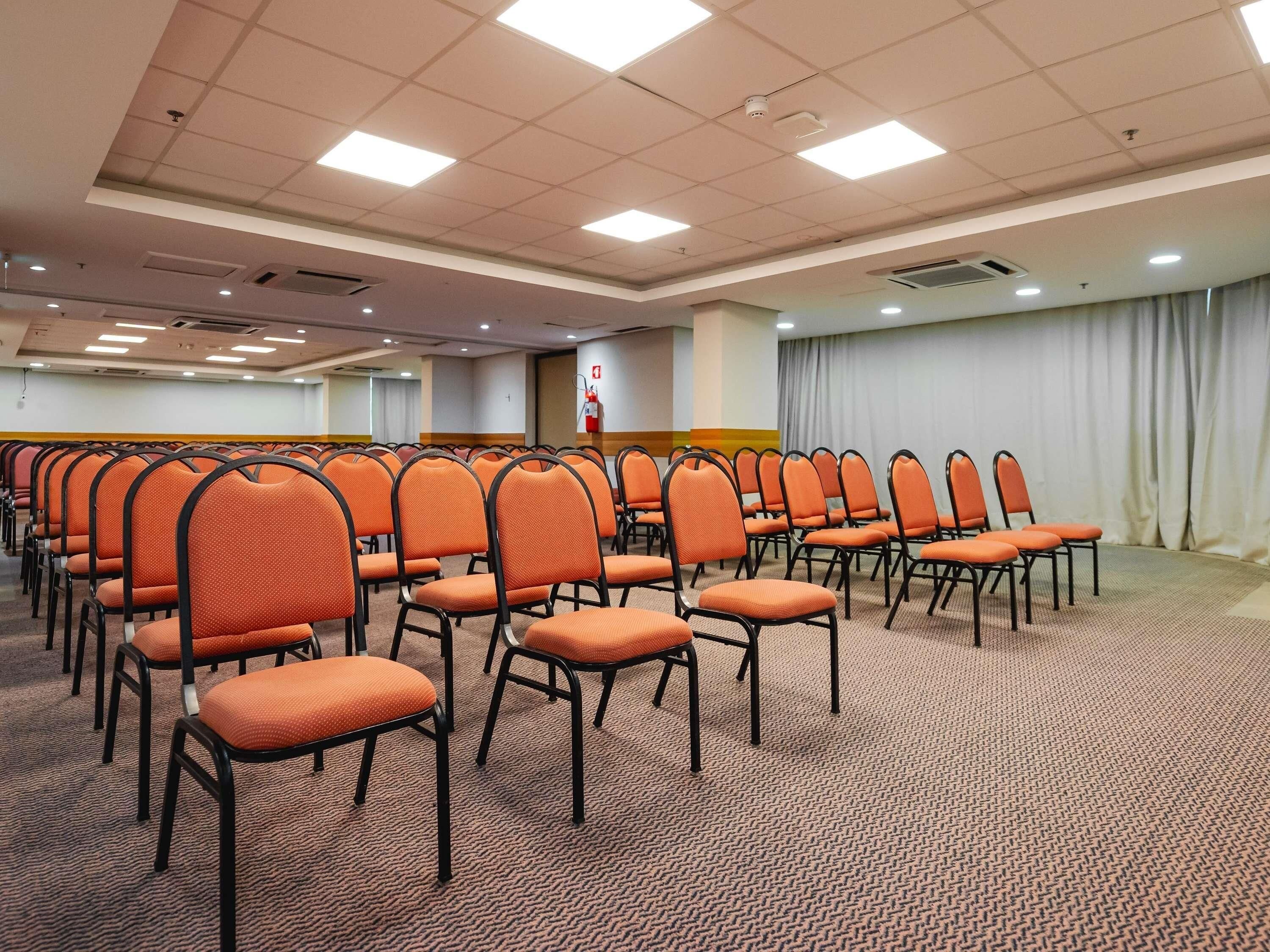 A conference room with rows of orange chairs, carpeted floor, and ceiling lights.