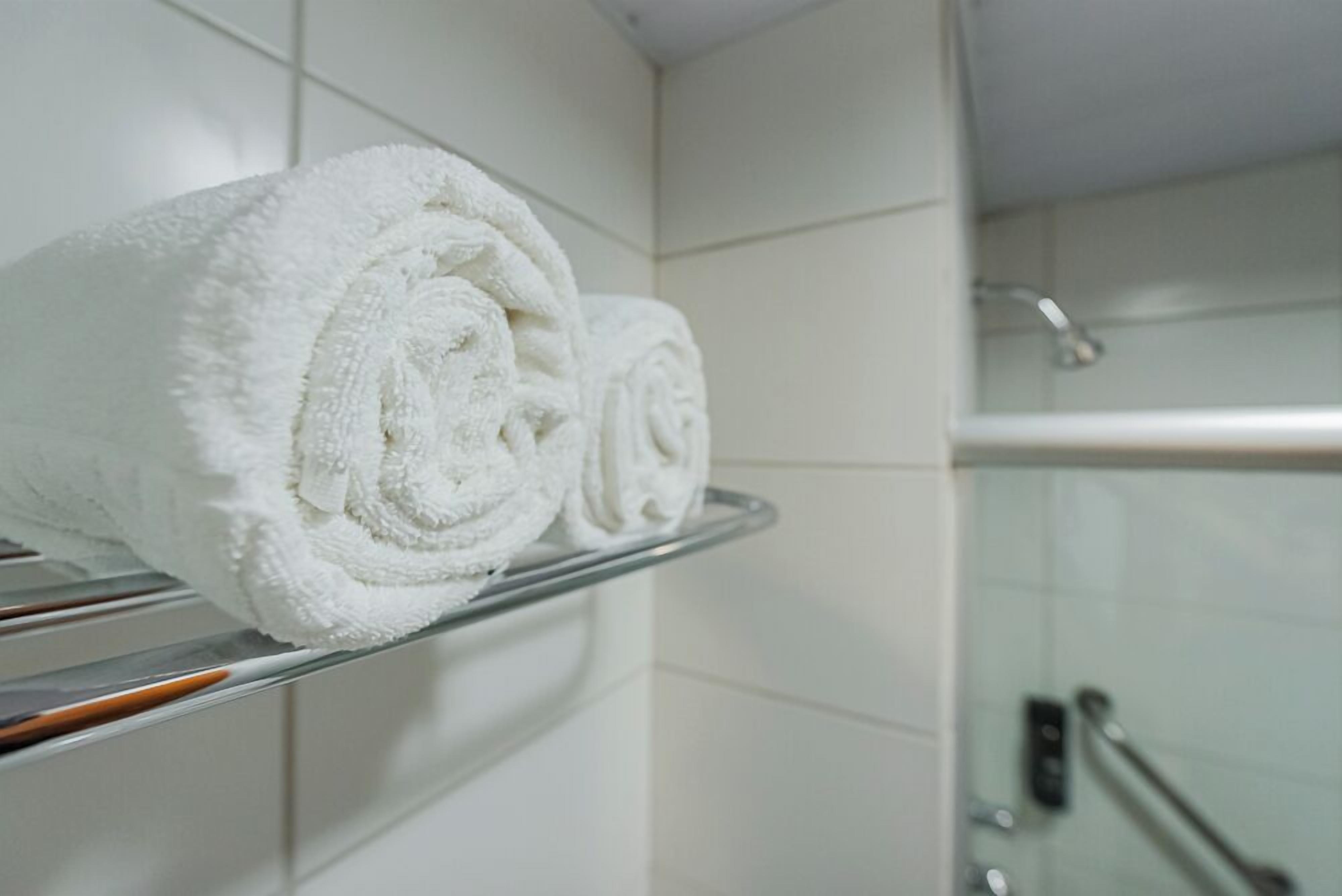 A bathroom with white tiled walls, a glass shower enclosure, and a metal towel rack with neatly rolled white towels.