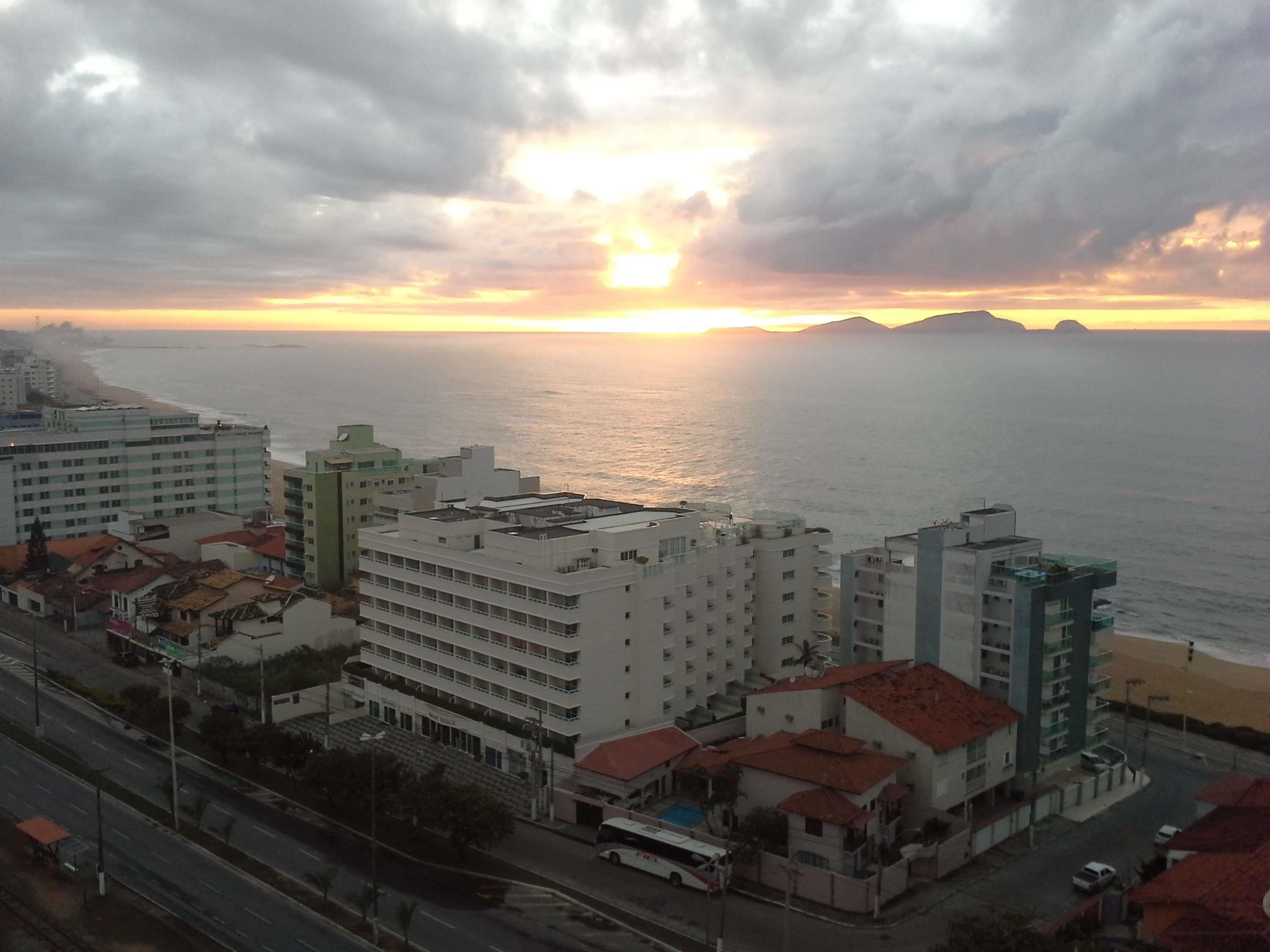 A coastal cityscape with buildings, a road, and the ocean at sunset.