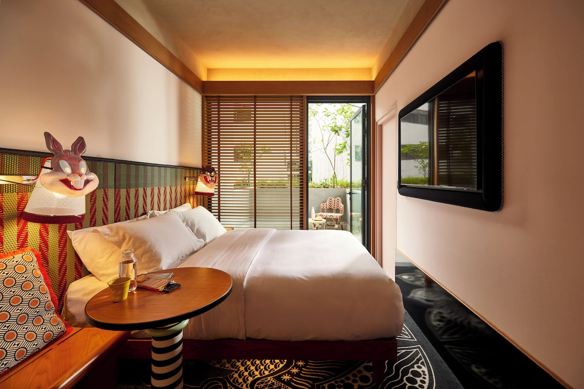 A hotel room with a bed, a small table, a lamp, and a large window.