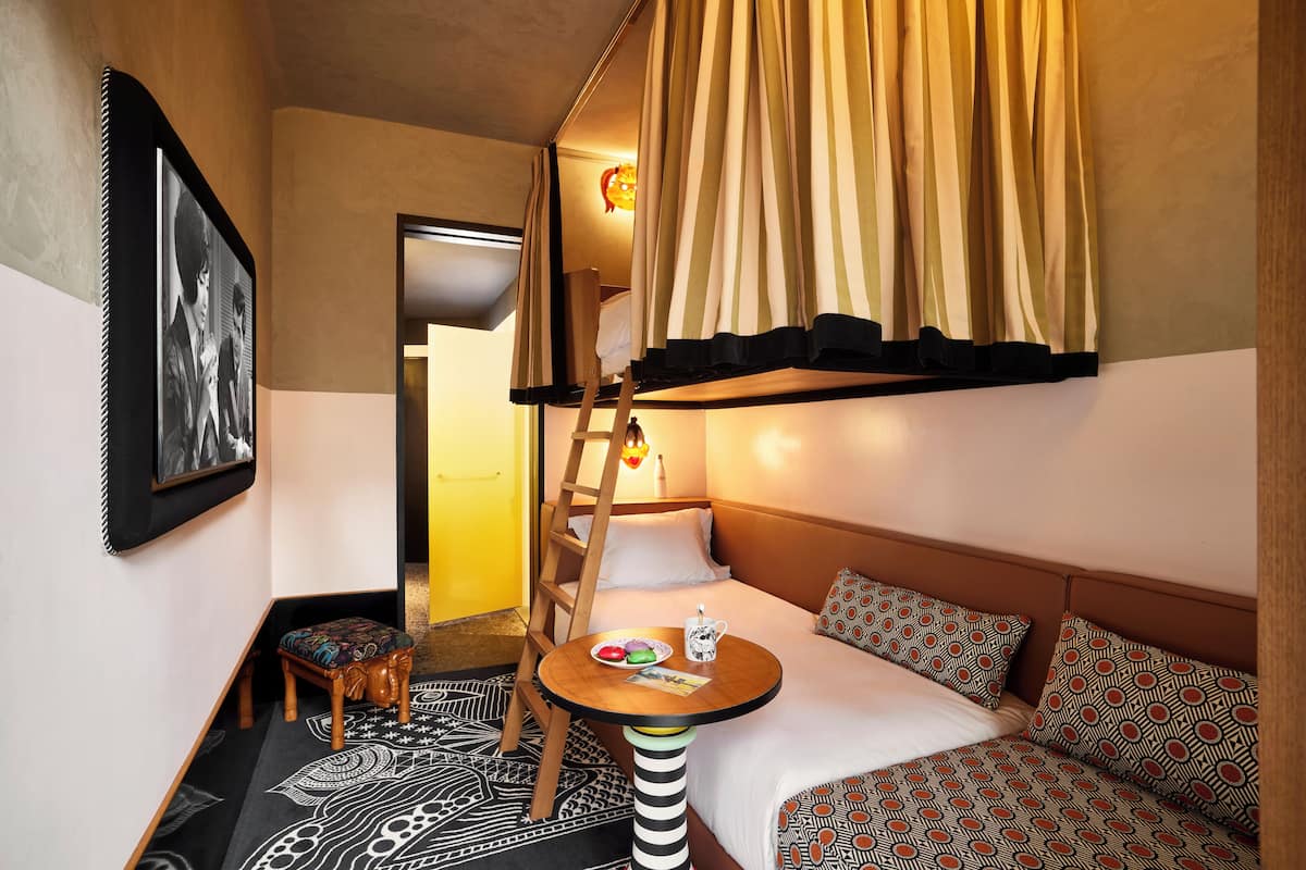 A compact hotel room with a bunk bed, a small table with a plate of food, a ladder, and a wall-mounted picture.