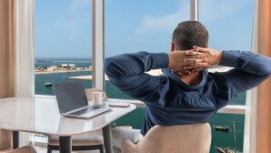 Um homem sentado à mesa com um laptop e uma xícara, com vista para um porto com barcos e edifícios.