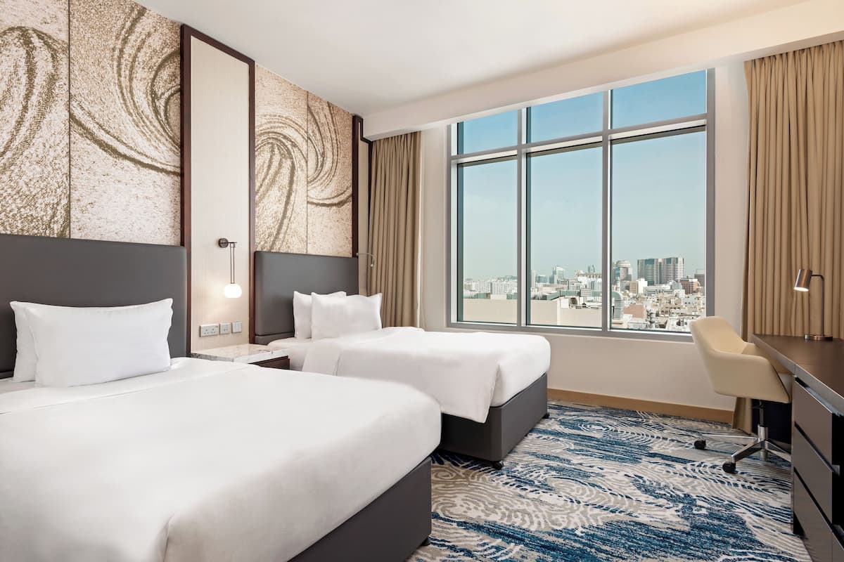 Une chambre d’hôtel avec deux lits, un bureau, une chaise, une grande fenêtre donnant sur la ville et une moquette à motifs.