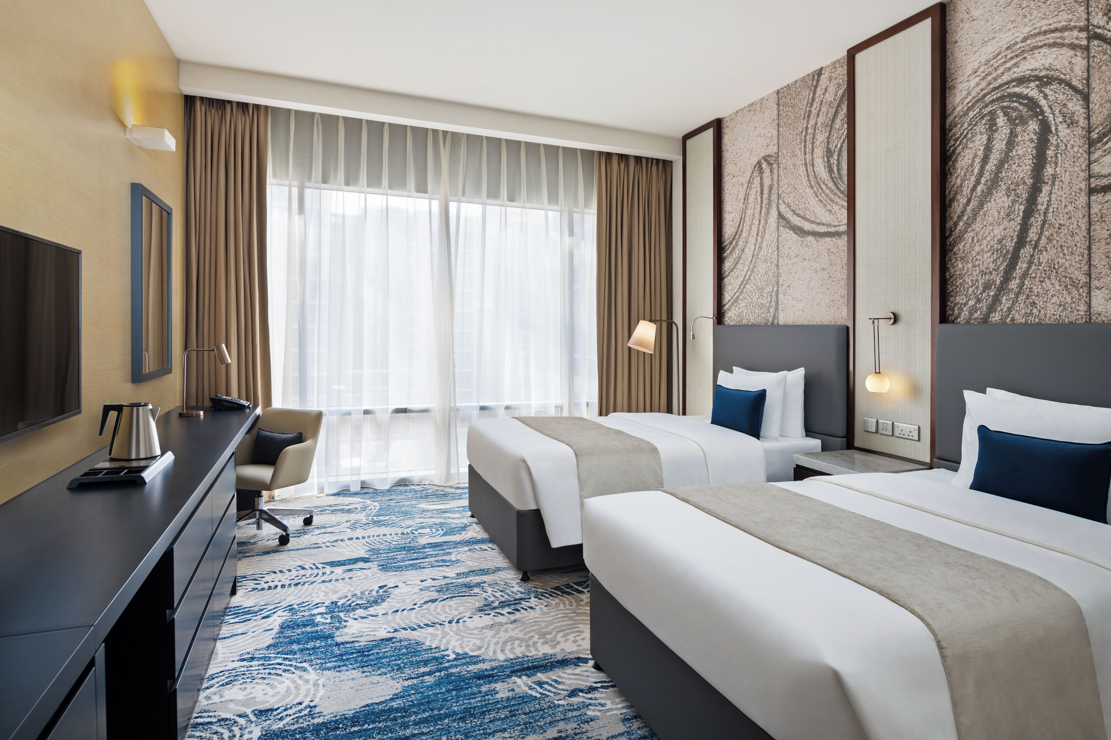 Habitación de hotel con dos camas, un escritorio, una silla, un televisor y un ventanal grande con cortinas.