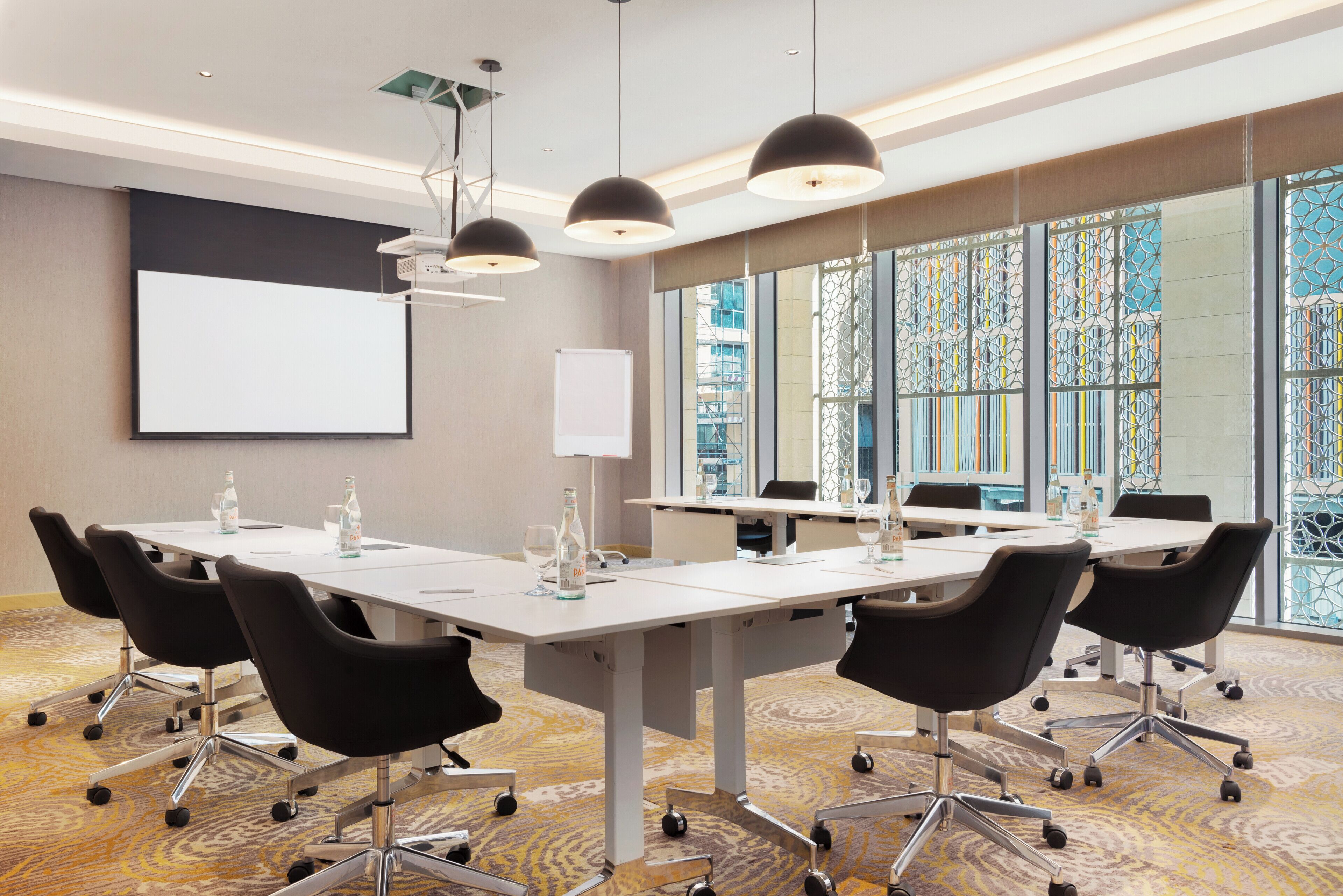 A conference room with a large table, chairs, a projector screen, and a view of a cityscape through large windows.