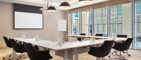 A conference room with a large table, chairs, a projector screen, and a view of a cityscape through large windows.