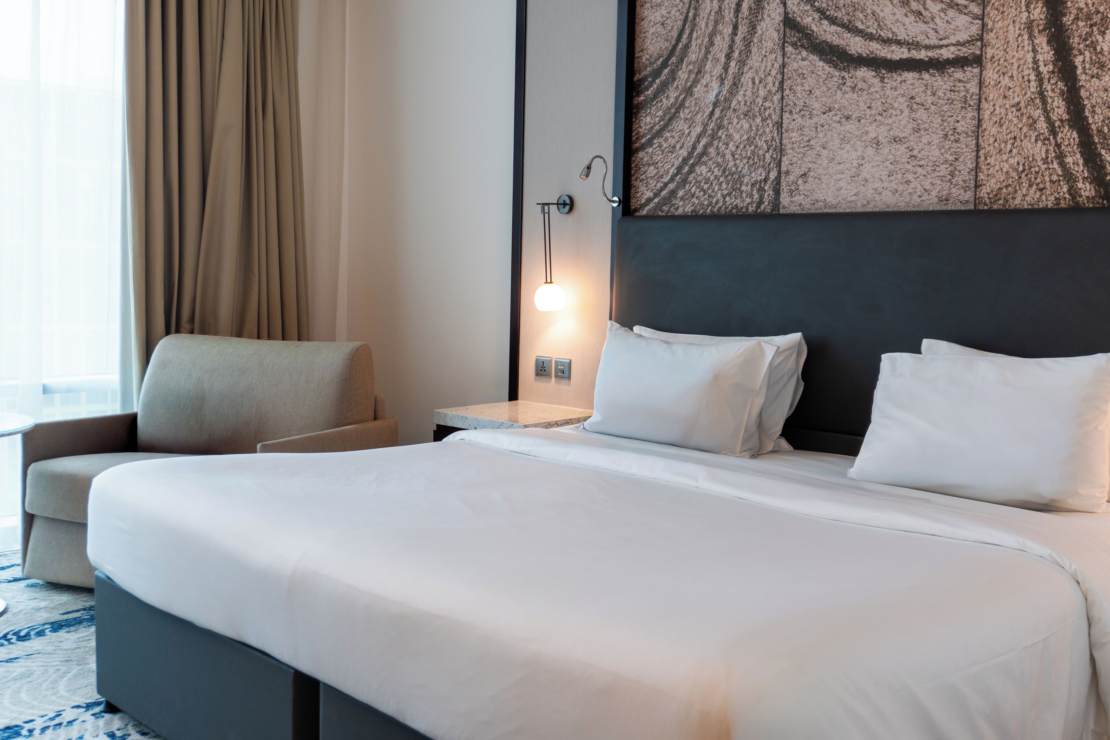 Una habitación de hotel con una cama grande, una mesita de noche, una lámpara, una silla y un cuadro en la pared.