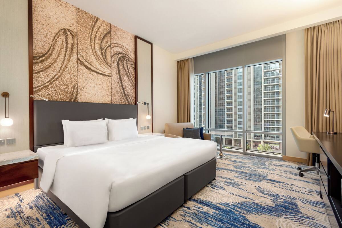 Une chambre d’hôtel avec un grand lit, un bureau, une chaise et une vue sur la ville grâce à de grandes fenêtres.