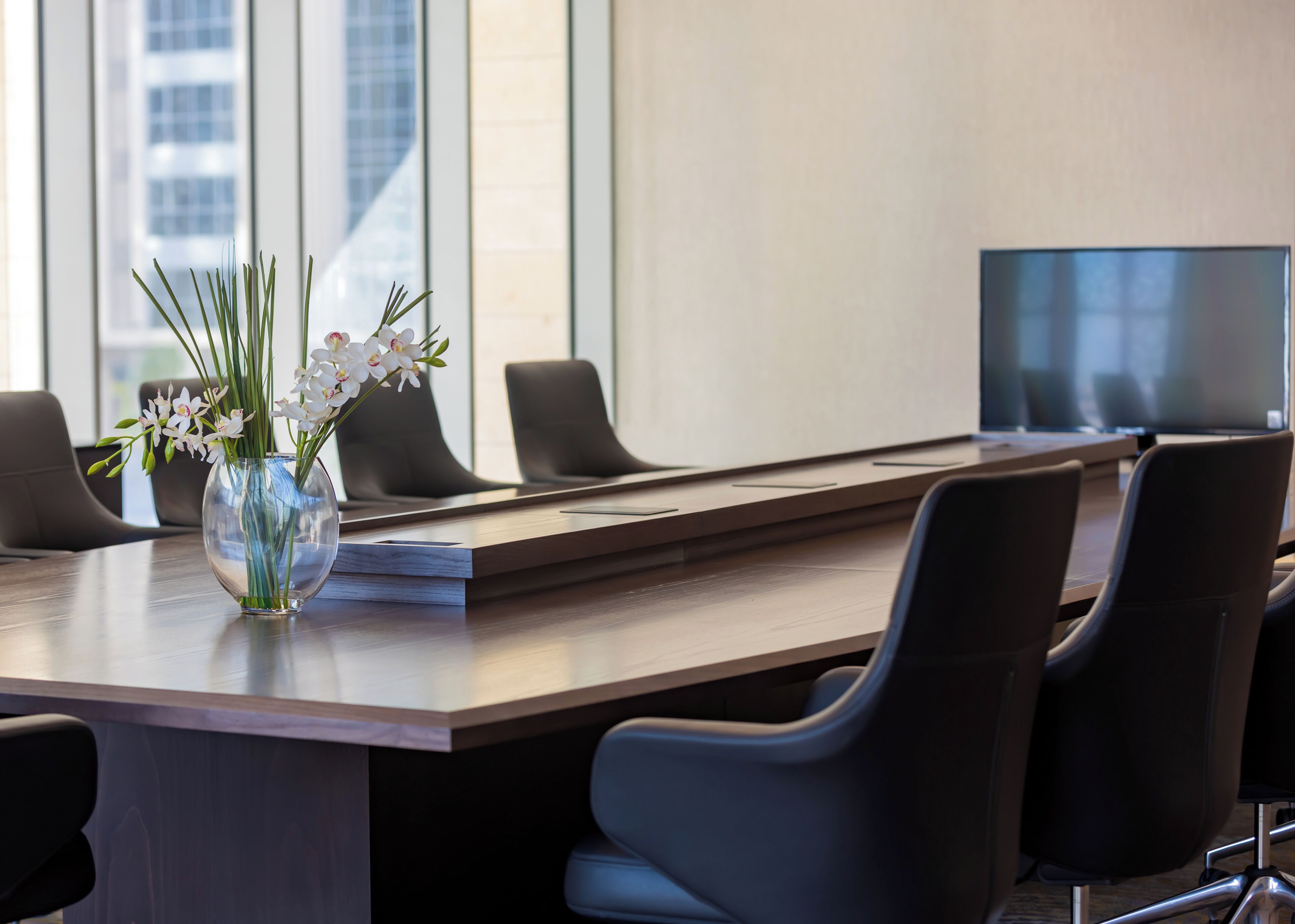 A conference room with a long table, six black chairs, a vase with white flowers, and a large screen.