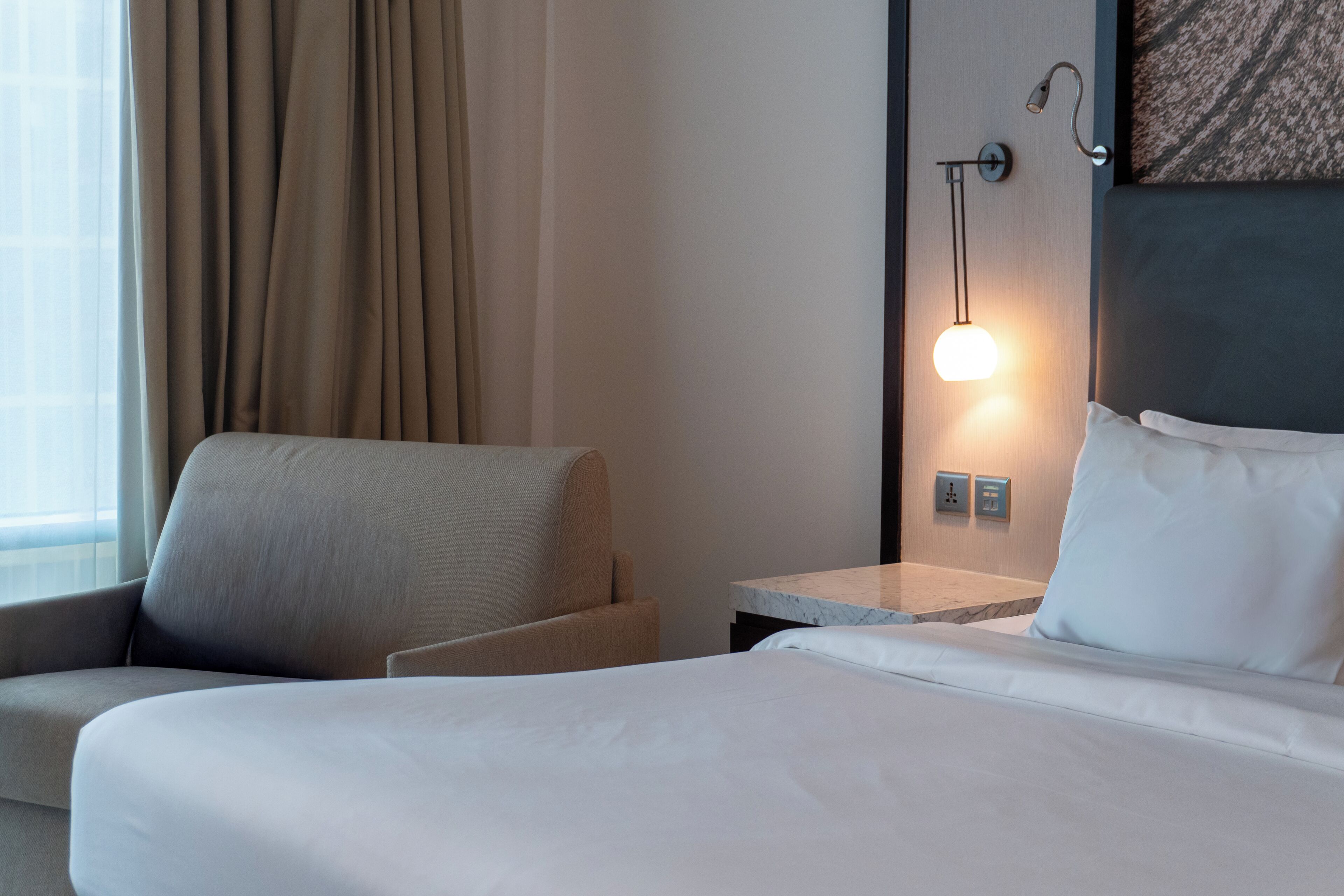 A hotel room with a bed, a chair, a nightstand, a wall-mounted lamp, and a window with curtains.