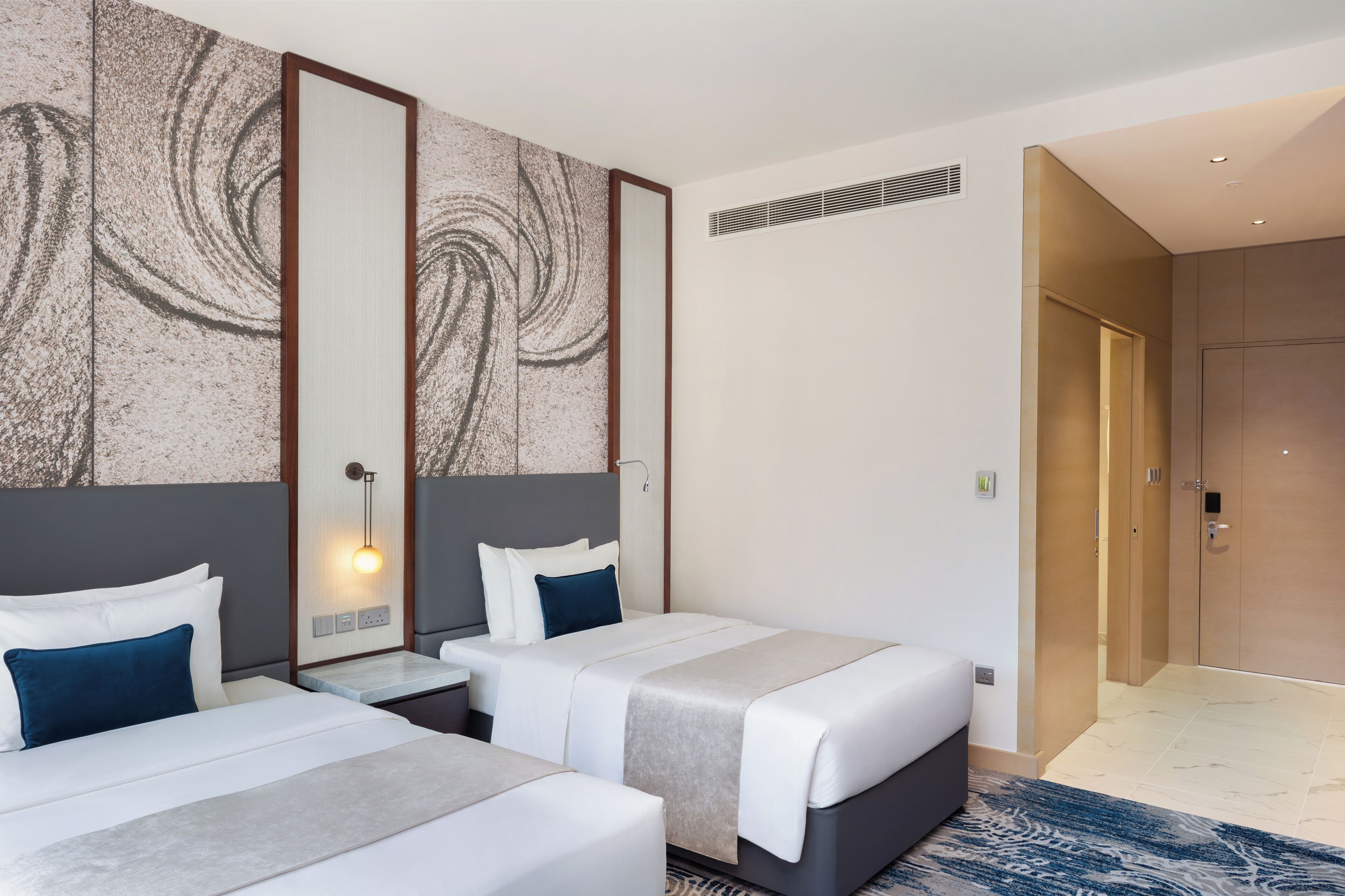 Une chambre d’hôtel avec deux lits, une grande œuvre d’art abstraite au mur, un luminaire moderne et une vue sur le couloir.