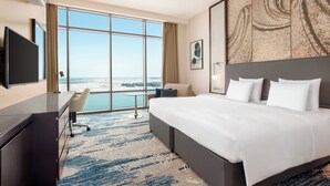Quarto de hotel com uma cama grande, uma escrivaninha, uma cadeira e uma televisão. Há uma janela ampla com vista para a praia.