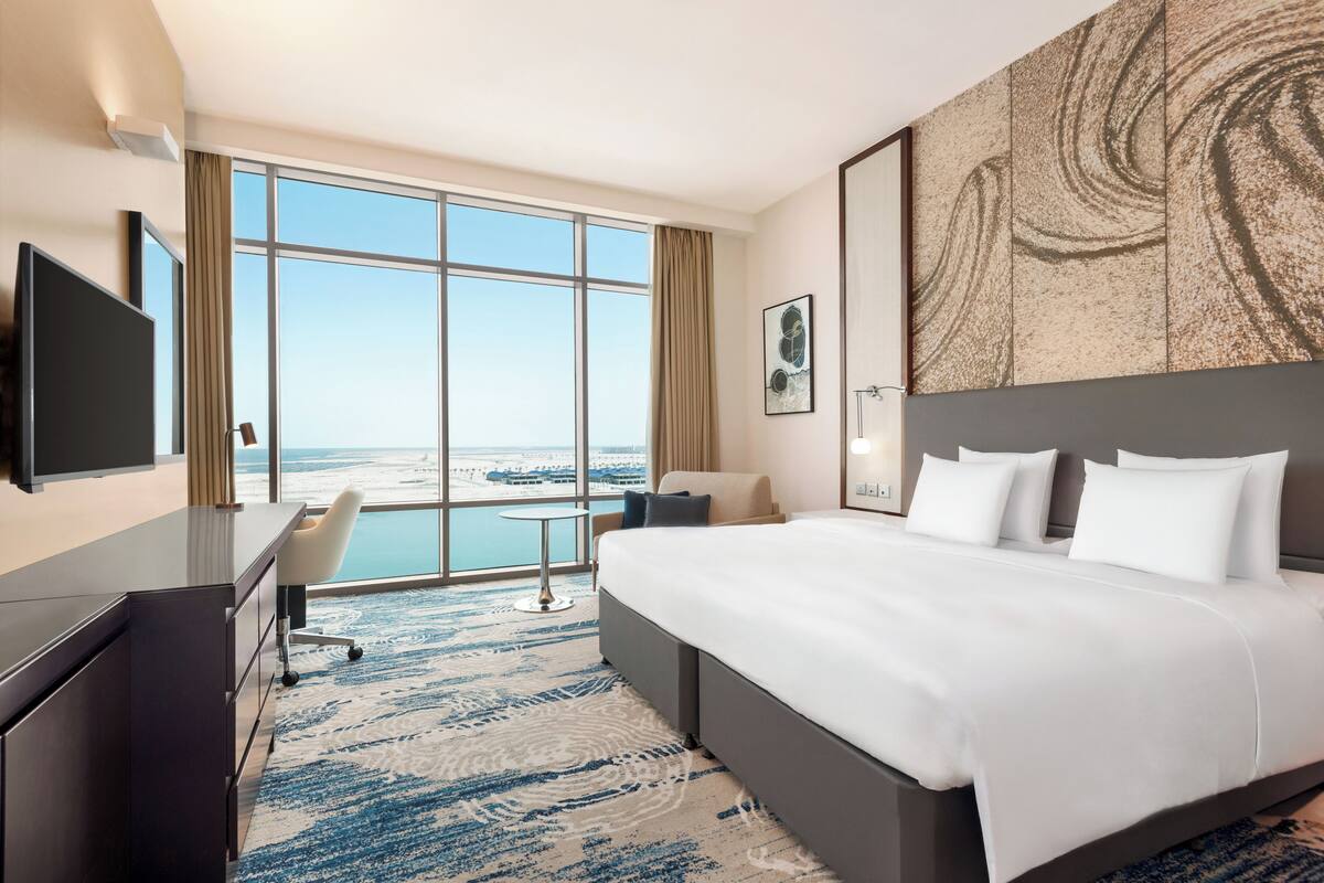 Une chambre d’hôtel avec un grand lit, un bureau, une chaise et une télévision. Une grande fenêtre offre une vue sur la plage.